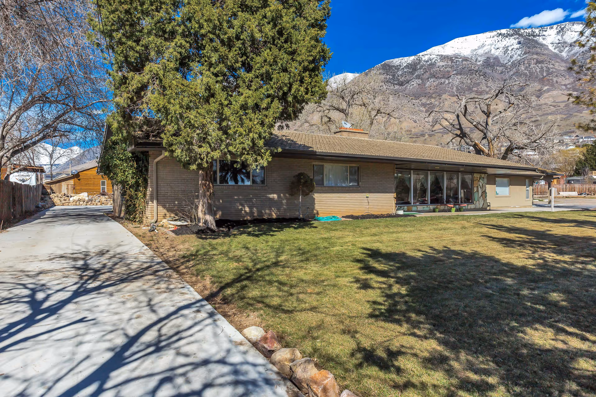 Single-story brick building with large windows, surrounded by a green lawn and trees, with a mountain covered in snow in the background under a clear blue sky.
