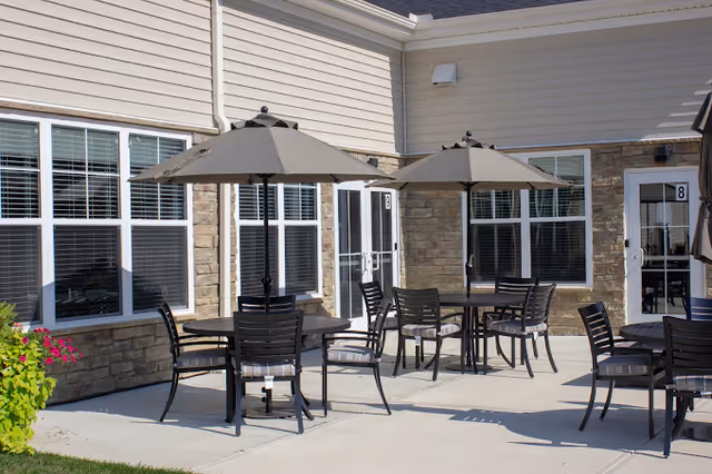 Outdoor patio area with round tables and black chairs, each table shaded by a large beige umbrella. The patio is adjacent to a building with stone and beige siding, featuring multiple windows and doors.