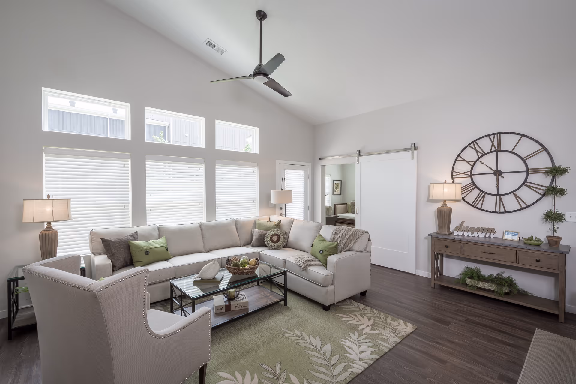 A bright and modern living room with a large beige sectional sofa adorned with green and brown pillows, a beige armchair, a glass coffee table with decorative items, a green patterned rug, and a wooden console table with a large round wall clock, lamps, and plants. The room features high ceilings with a ceiling fan and multiple windows with white blinds.