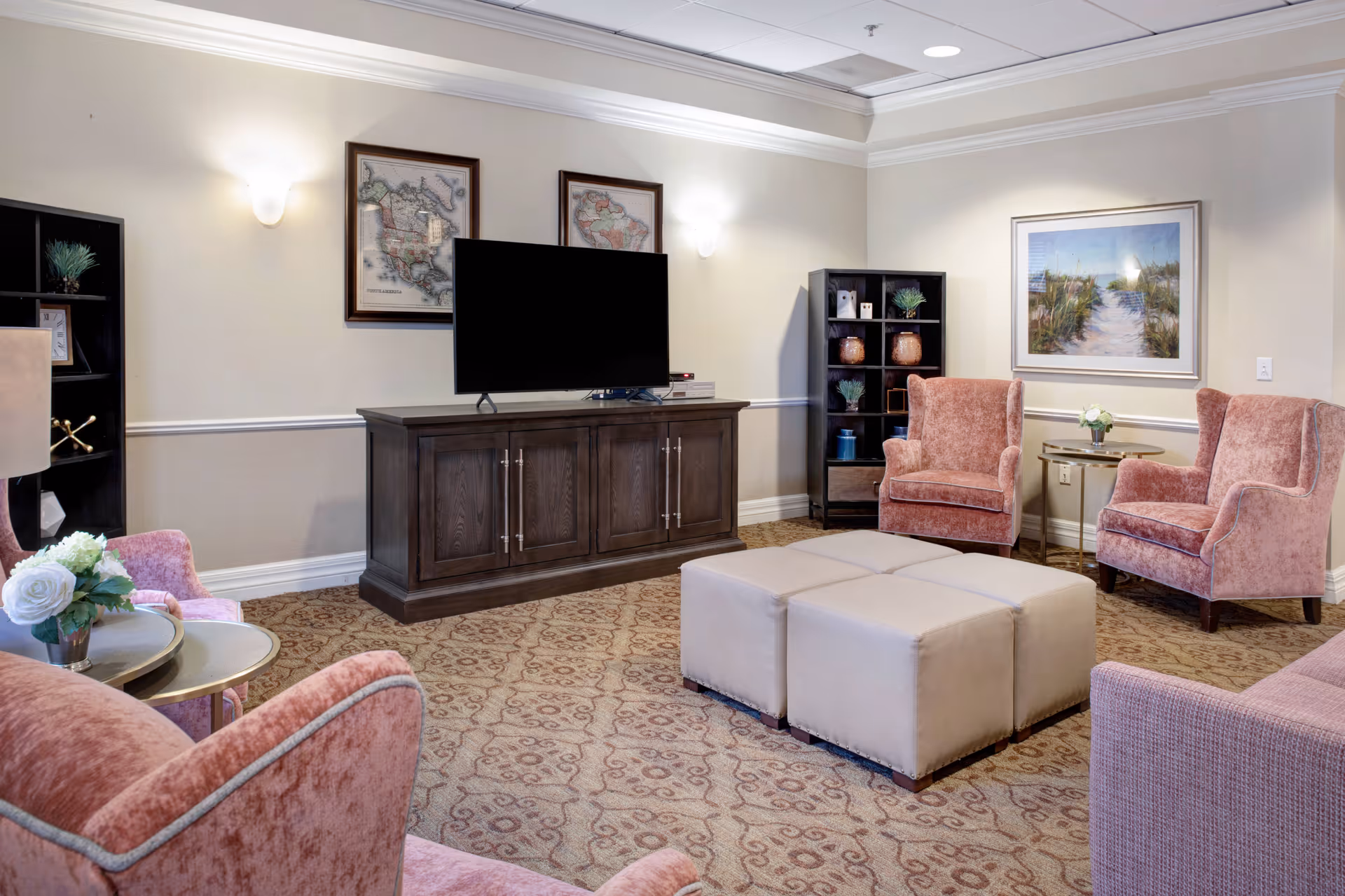 A cozy living room area with pink upholstered armchairs and a matching sofa arranged around a set of four beige ottomans. A dark wooden TV stand holds a flat-screen television, flanked by two black shelving units with decorative items. The walls are light-colored with framed artwork and soft wall sconces providing lighting.