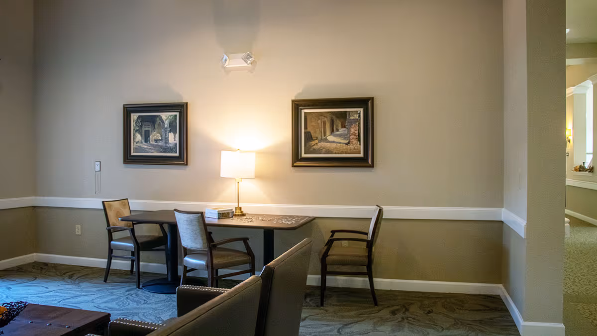 A cozy lobby area featuring a small table with two chairs, a lamp, and framed artwork on the walls. The decor includes soft lighting and a patterned carpet.