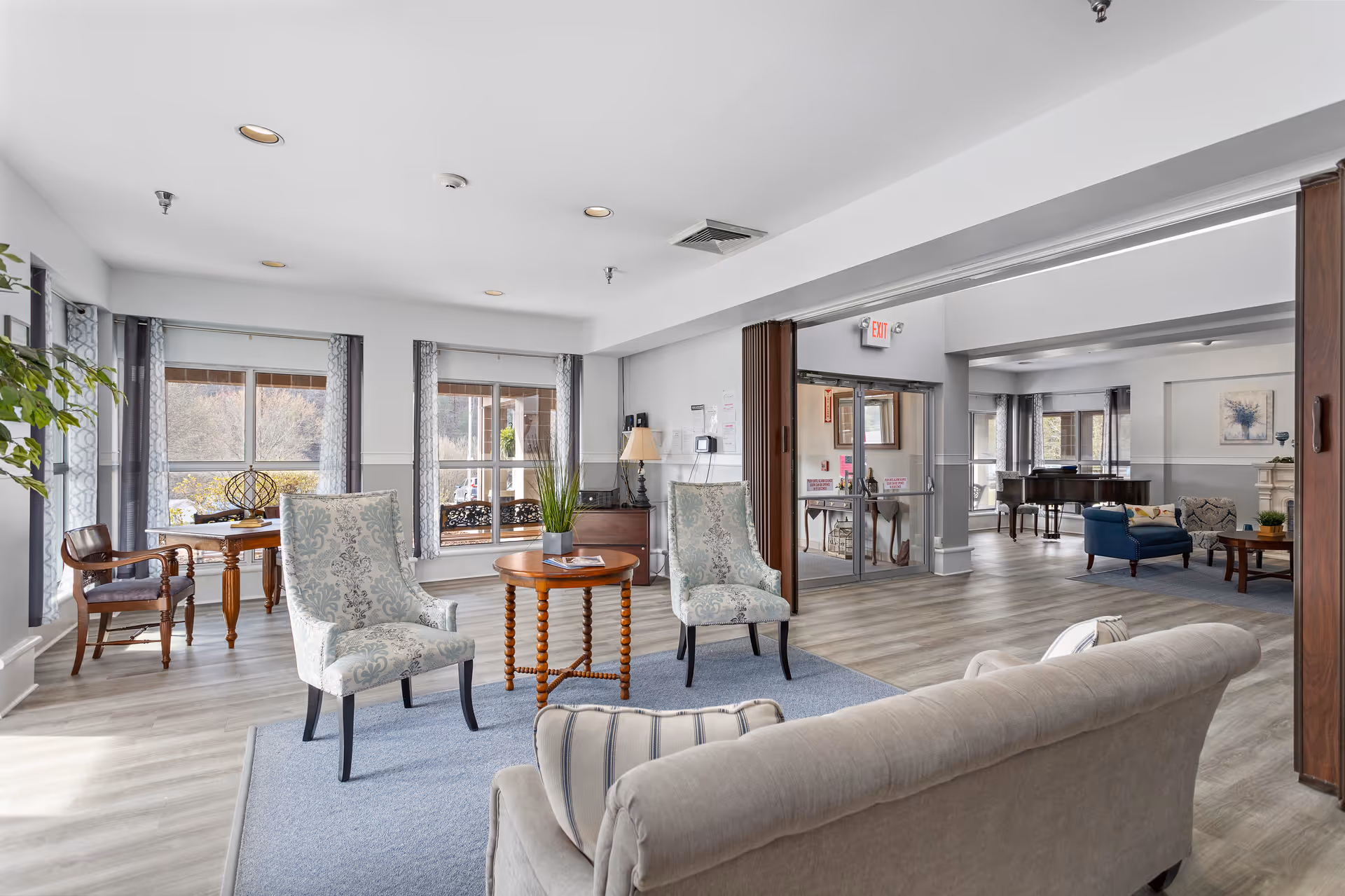 A bright and spacious senior living facility common area with large windows letting in natural light. The room features comfortable upholstered chairs and a sofa arranged around a wooden coffee table on a blue rug. In the background, there is a grand piano and additional seating, with light-colored walls and wood flooring throughout.