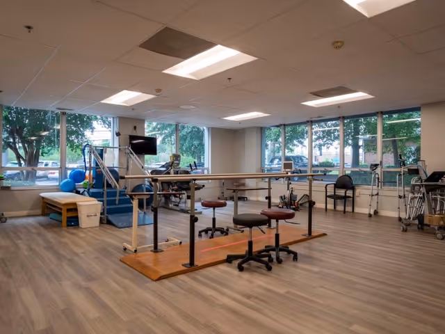 A spacious rehabilitation room with large windows showing trees outside. The room features parallel bars for walking exercises, three rolling stools, exercise equipment including a treadmill and therapy balls, and chairs along the walls. The floor is wood-patterned, and the ceiling has recessed lighting.