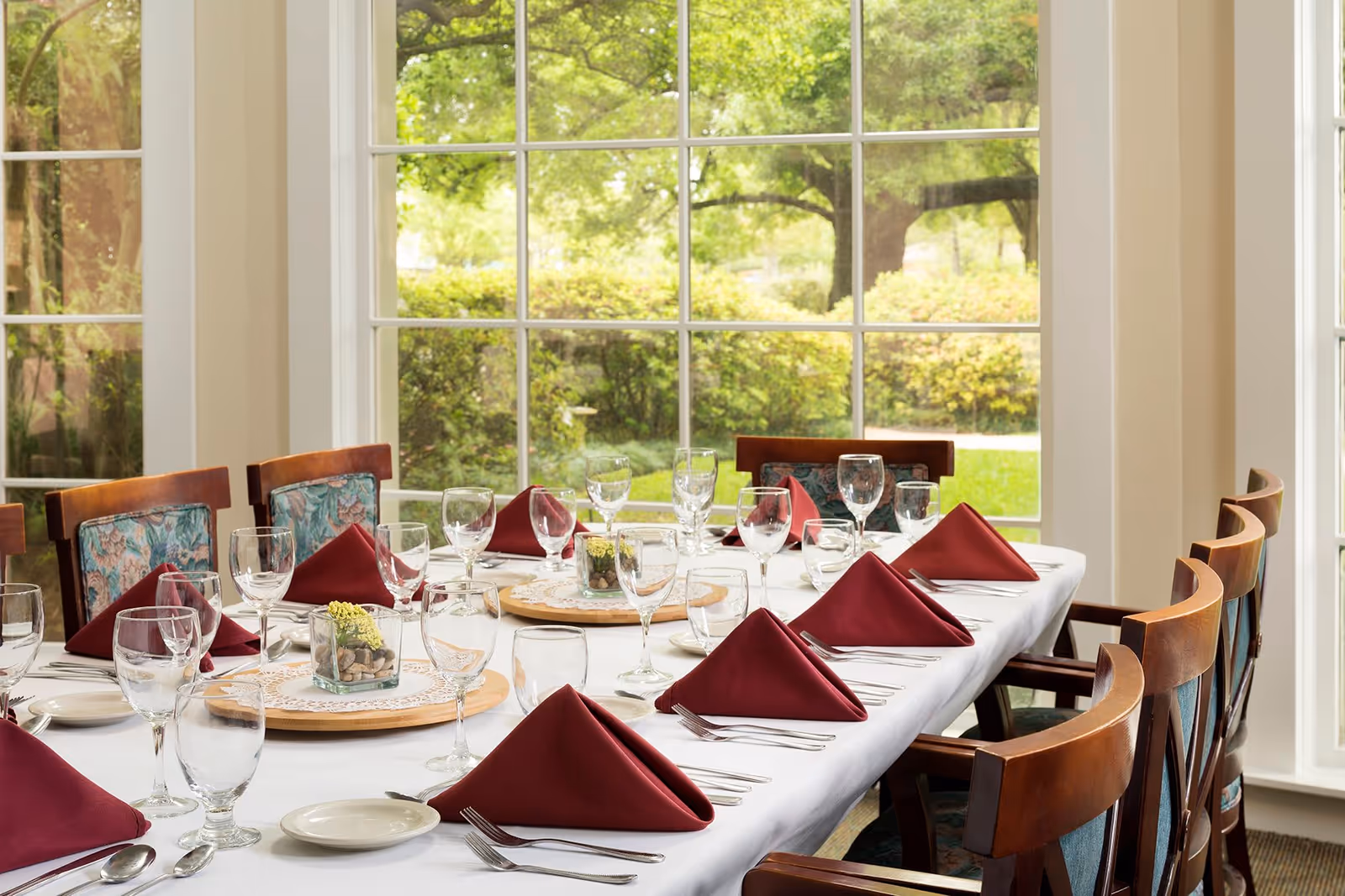 A dining table set for a meal with white tablecloth, burgundy folded napkins, multiple glasses, plates, and silverware. The table is surrounded by wooden chairs with patterned cushions. Large windows in the background show a green outdoor garden area.