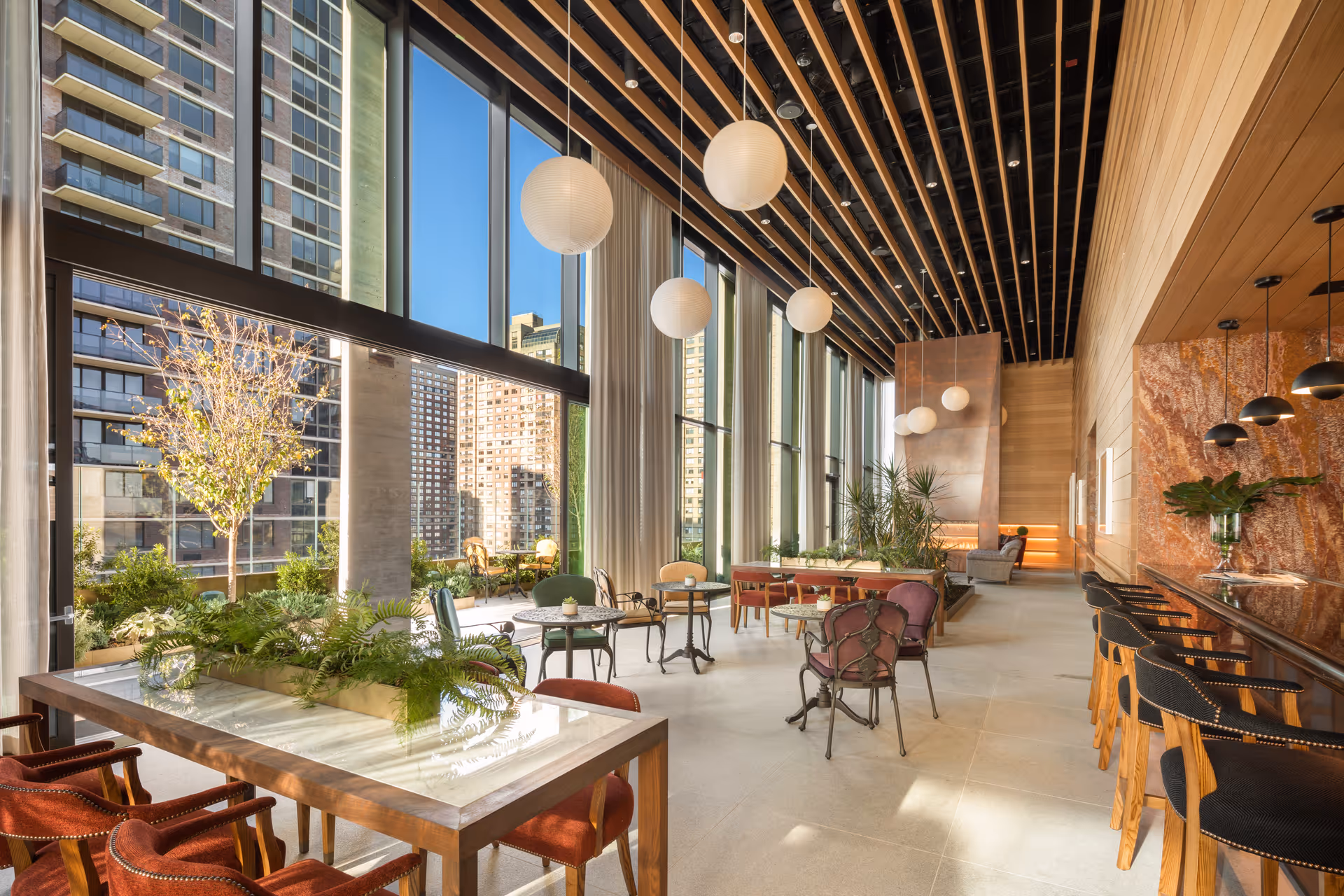 A spacious, modern interior lounge area with large floor-to-ceiling windows letting in natural light. The room features a variety of seating options including red cushioned chairs around a wooden table with plants, small round tables with chairs, and a bar area with black and wooden stools. The ceiling has a wooden slat design with hanging round white pendant lights. Outside the windows, tall buildings and greenery are visible.