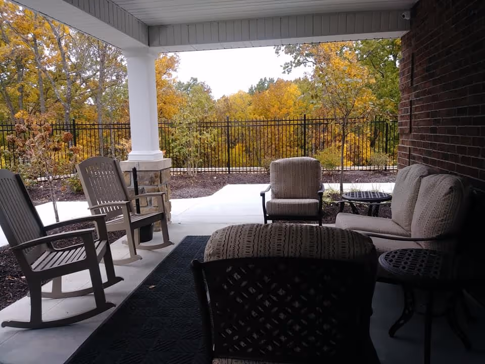 Covered outdoor patio area with cushioned chairs, rocking chairs, and small tables. The patio overlooks a fenced garden area with trees showing autumn foliage.
