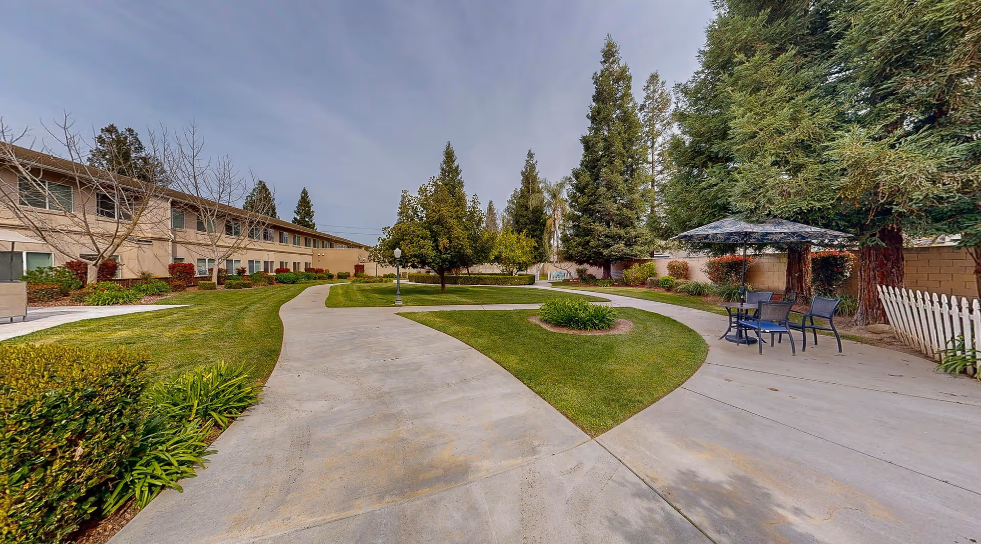 Outdoor courtyard area at Cogir of Cedar Creek featuring a winding concrete pathway surrounded by well-maintained grass, trees, and shrubs. There is a seating area with a table and chairs under a large umbrella on the right side, and a two-story building with multiple windows is visible on the left.