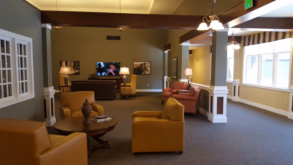 A cozy living room area in a senior living facility with mustard yellow armchairs around a round wooden coffee table, a brown couch facing a wall-mounted TV, and additional seating with a red armchair and side lamps. The room has beige walls, carpeted floors, large windows letting in natural light, and decorative framed pictures on the walls.