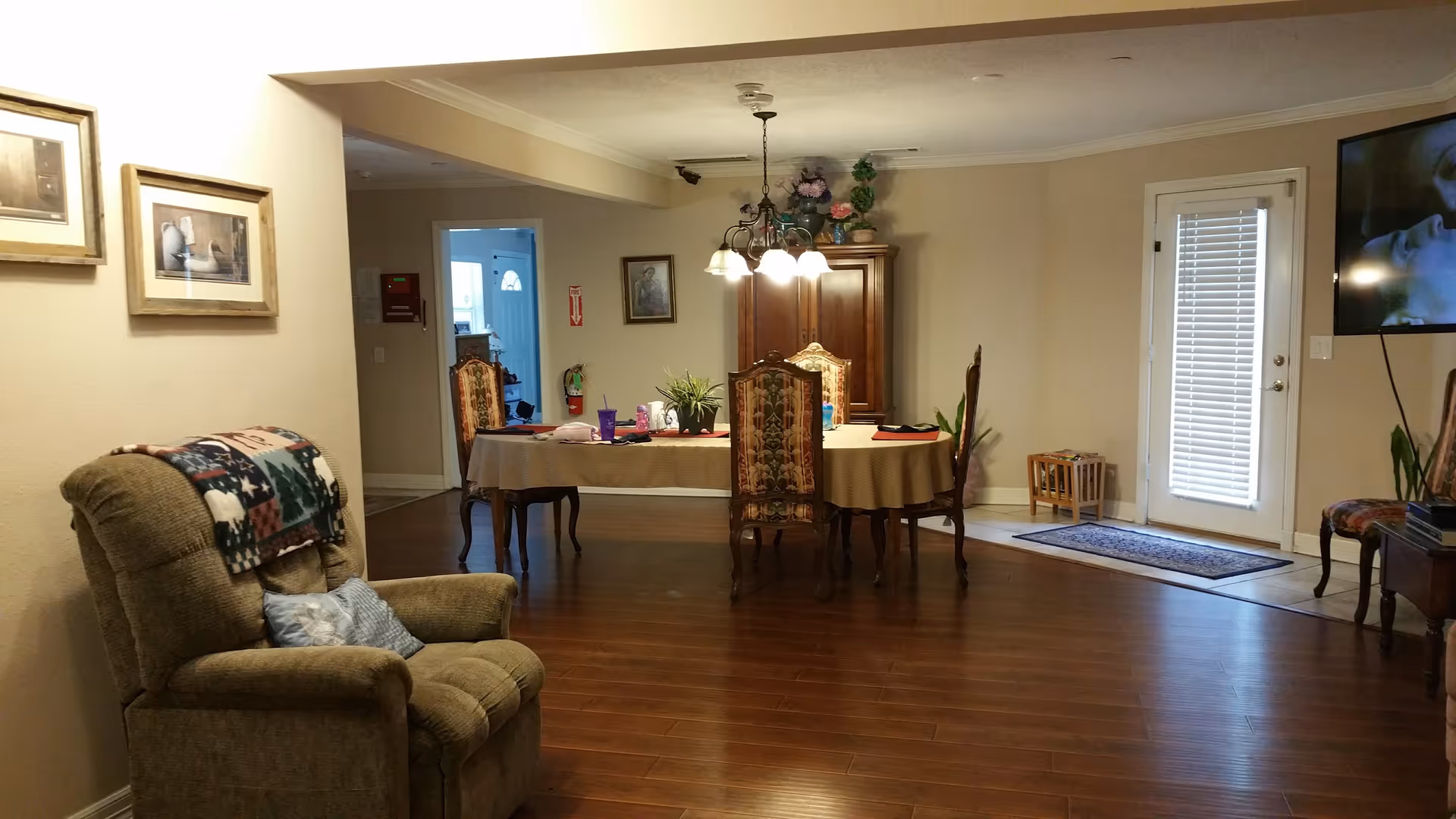 Open living and dining area with a recliner in the foreground, a dining table under a chandelier in the middle, and a TV and door on the right.