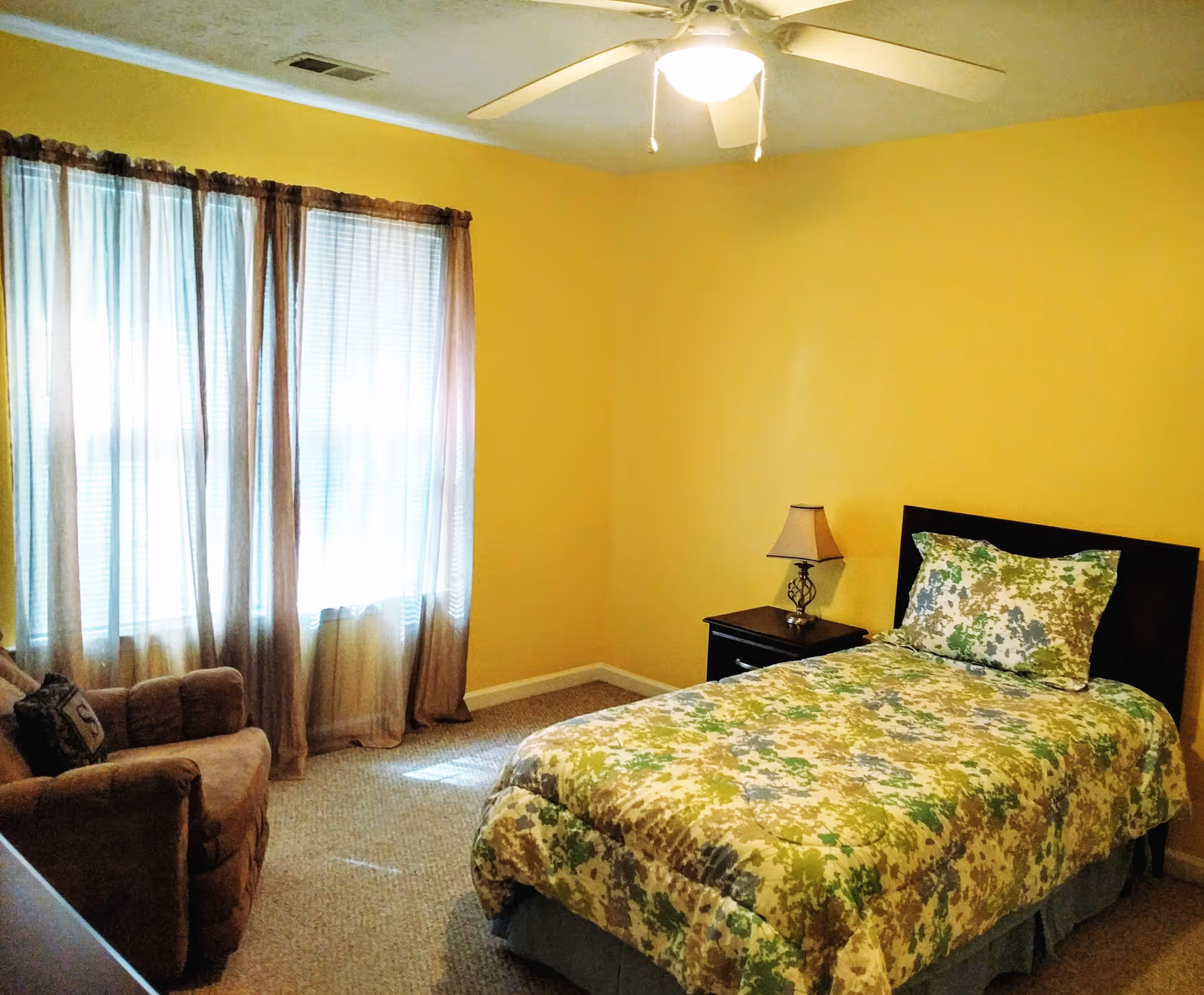 A bright bedroom with a single bed, nightstand and lamp, an armchair, and large curtained windows.
