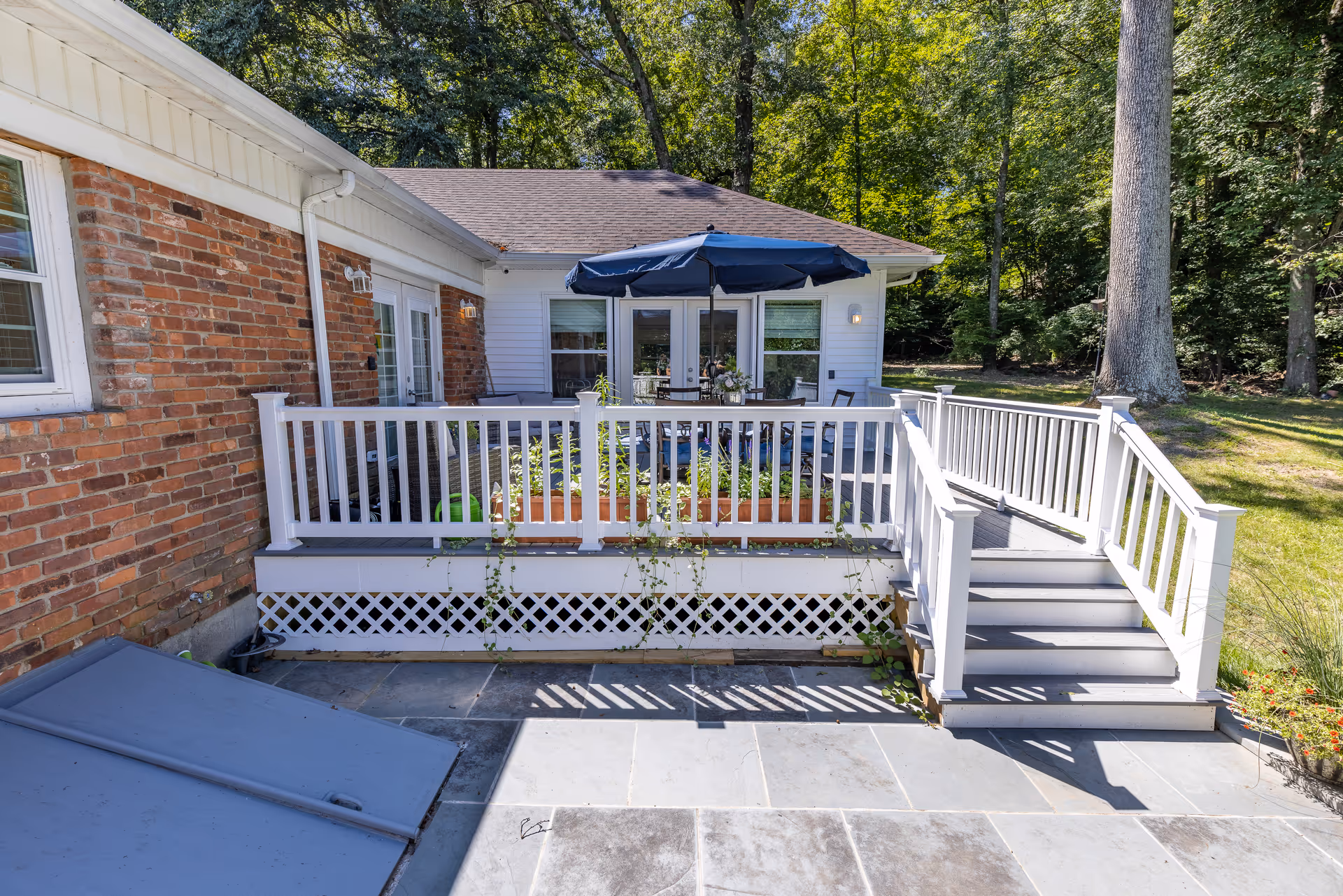 Backyard deck with white railing and stairs leading to a patio table under a blue umbrella beside a house with brick and siding, surrounded by trees.