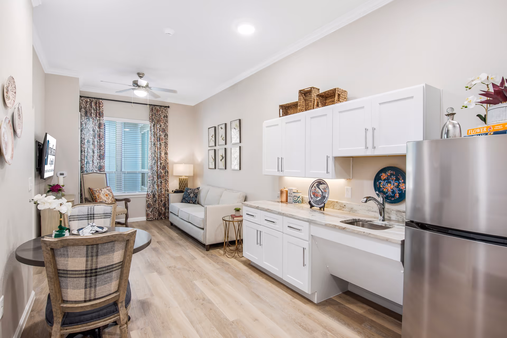 A bright and cozy senior living apartment interior featuring a small kitchen area with white cabinets, a marble countertop, and a stainless steel refrigerator. Adjacent to the kitchen is a living area with a beige sofa, a side table with a lamp, and a window with patterned curtains. A small round dining table with two plaid upholstered chairs is positioned near the kitchen. The room has light wood flooring and neutral-colored walls decorated with framed artwork and decorative plates.