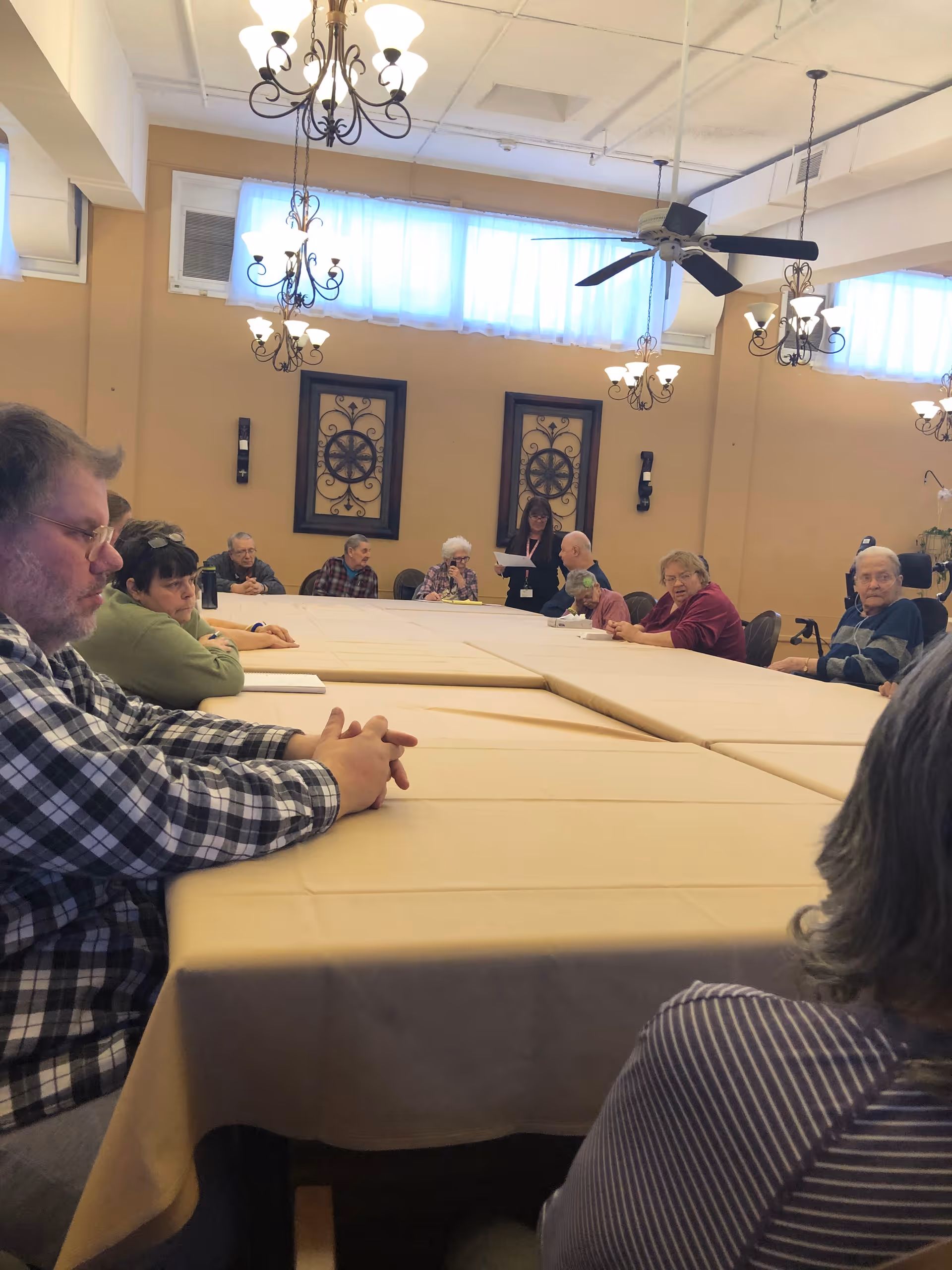 Residents seated around a large table in a communal dining/activity room with chandeliers and wall decor.