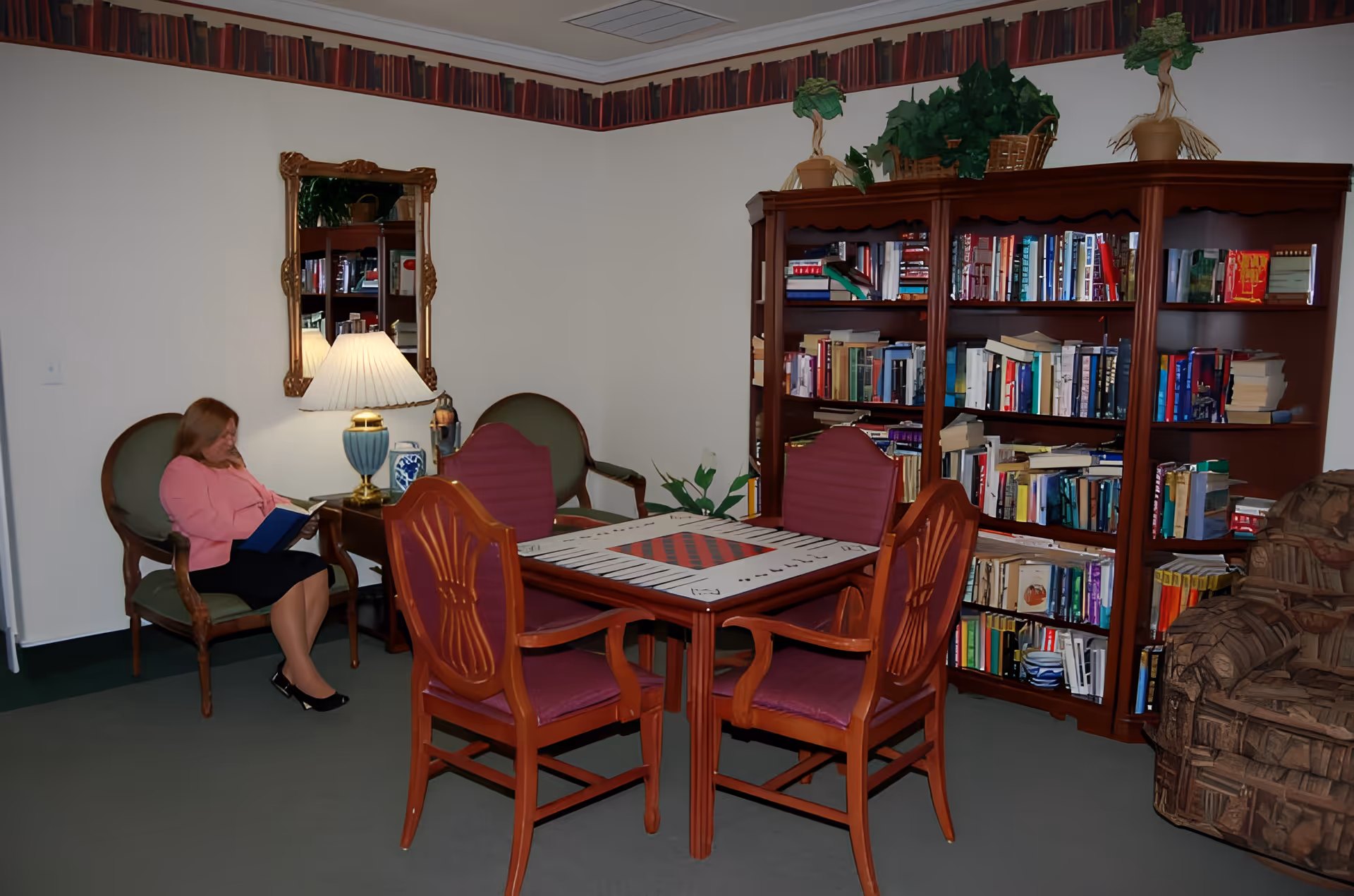 A cozy reading and game area in an assisted living facility featuring a woman sitting on a green armchair reading a book. There is a wooden table with a checkerboard pattern on top surrounded by four wooden chairs with red cushions. Behind the table is a large wooden bookshelf filled with books and decorated with potted plants on top. A lamp on a small side table next to the woman provides warm lighting, and a mirror hangs on the wall above the lamp. The room has a carpeted floor and a wallpaper border with a book pattern near the ceiling.