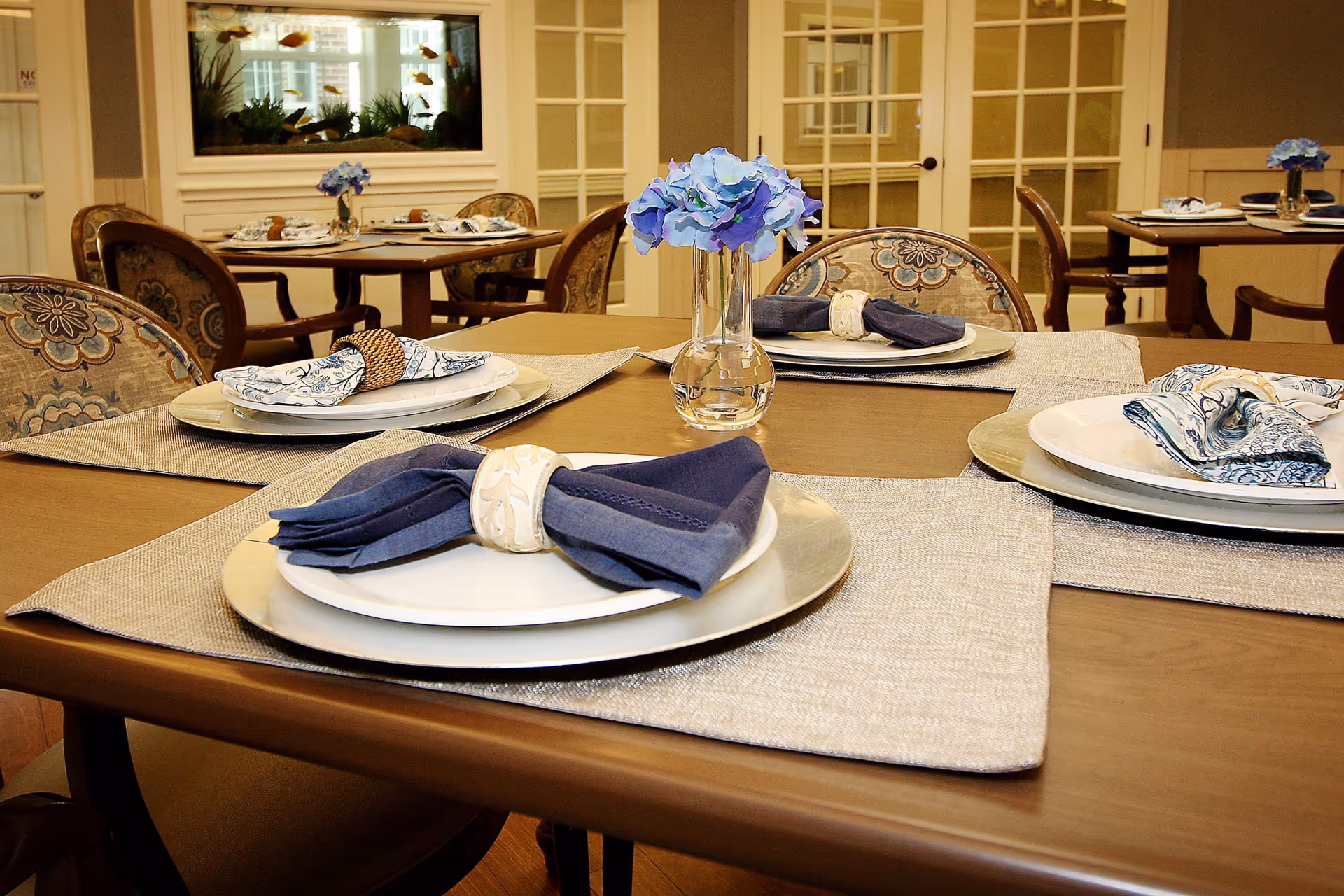 A dining room with wooden tables set with white plates, cloth napkins in napkin rings, and placemats. Each table has a small vase with blue flowers. The chairs have patterned upholstery. In the background, there is a large fish tank built into the wall and glass-paneled double doors.