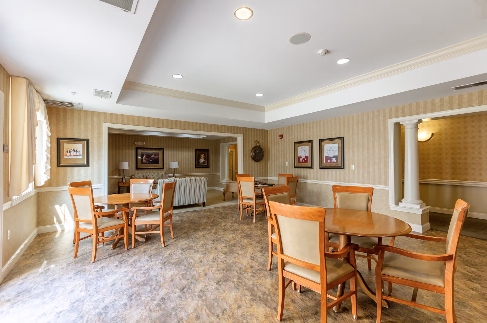 A well-lit common area in a senior living facility featuring multiple wooden tables with cushioned chairs arranged around them. The room has beige patterned wallpaper, a light-colored ceiling with recessed lighting, and large windows with curtains allowing natural light to enter. In the background, there is a seating area with striped sofas, side tables, and framed artwork on the walls.