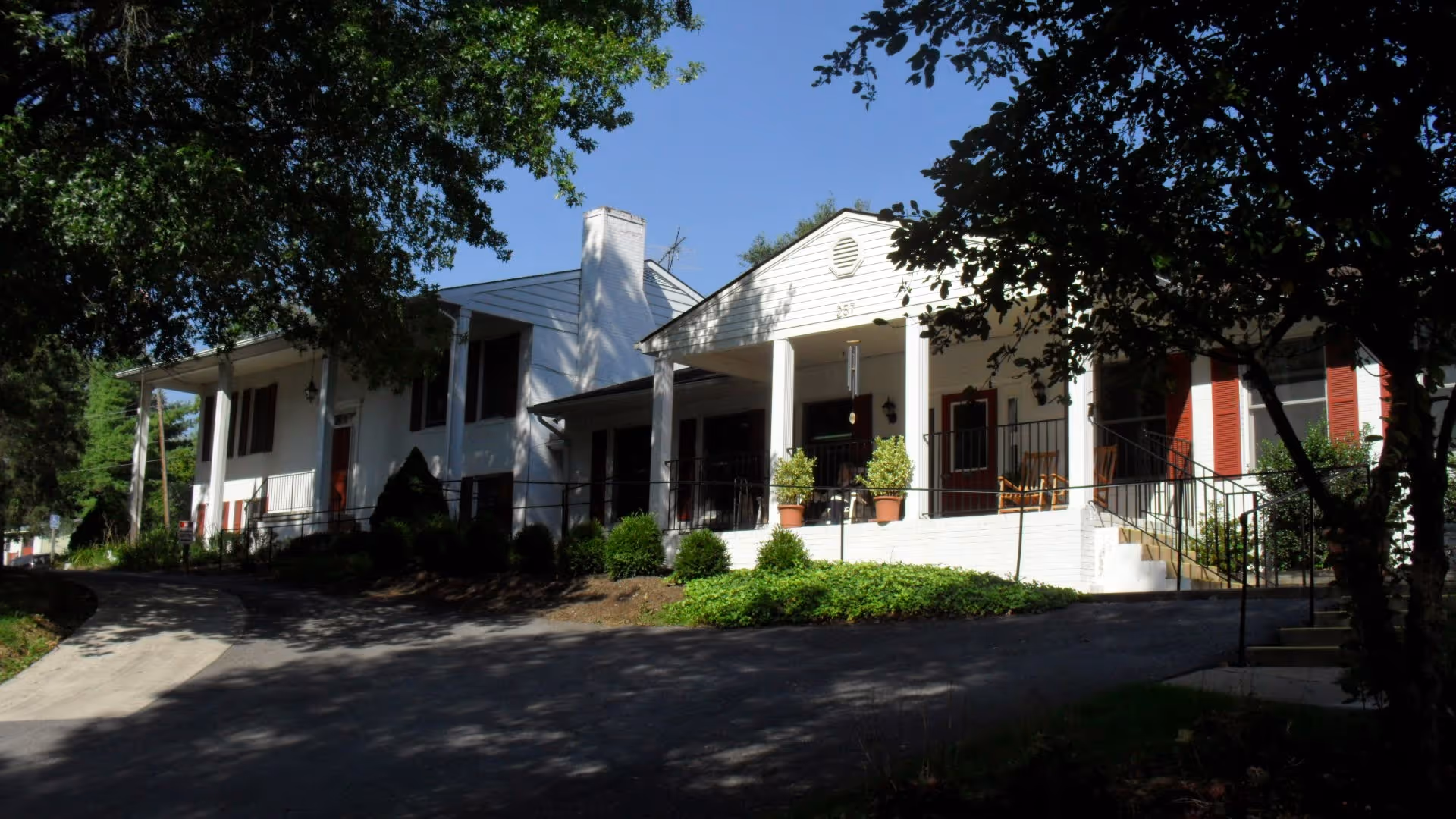 Exterior view of Dutch Haven Assisted Living facility showing a white building with a covered porch, red doors, and windows with red shutters. The building is surrounded by trees and greenery with a paved driveway leading up to the entrance.