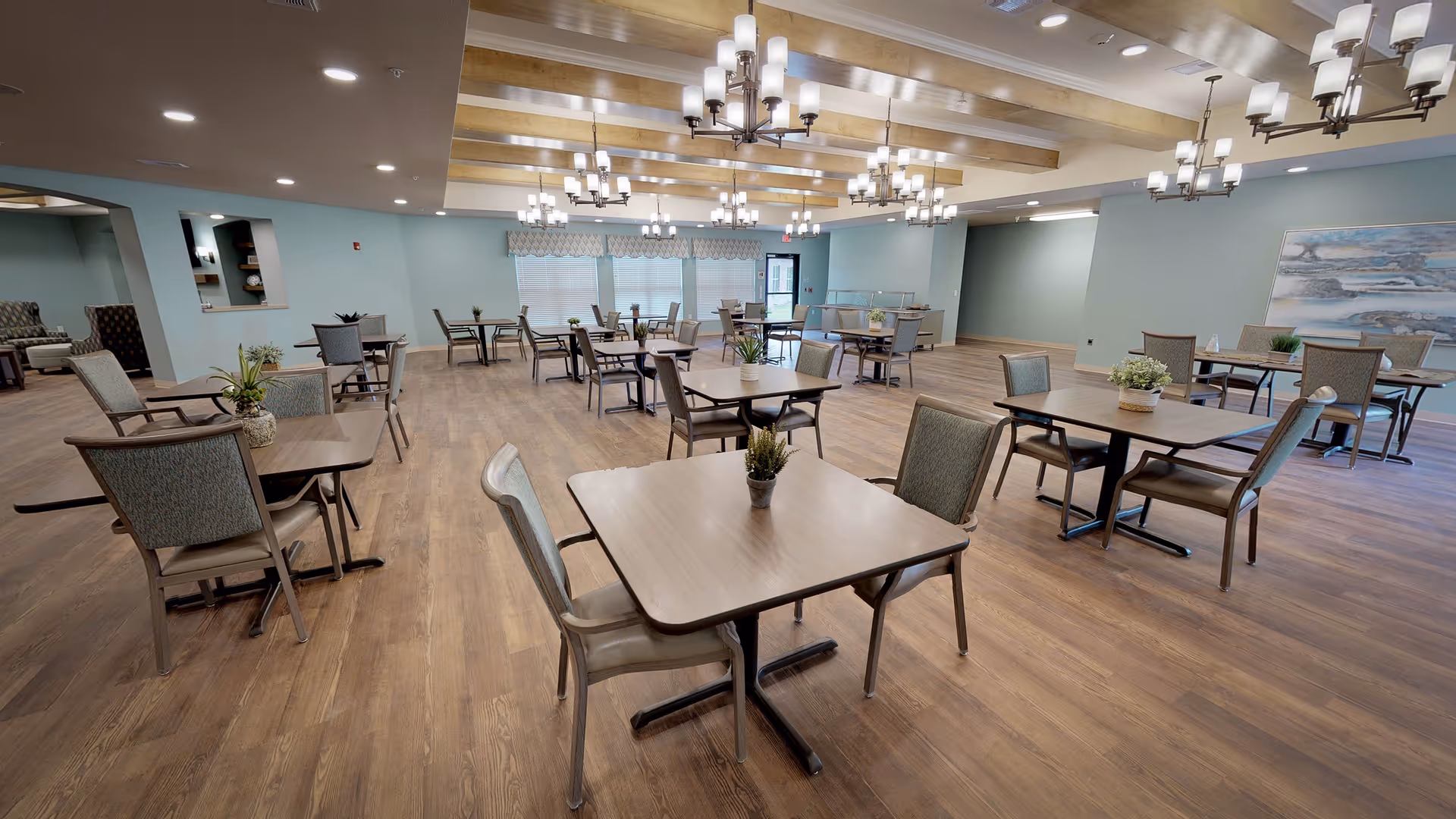 Large communal dining room with multiple square tables and chairs, wood flooring, soft teal walls, and chandeliers.
