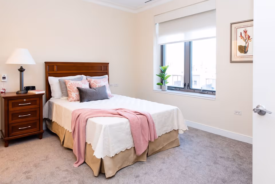 A neatly made bed with white bedding, pink and gray decorative pillows, and a pink throw blanket in a bright bedroom. There is a wooden nightstand with a lamp and a book next to the bed. A window with a white roller shade lets in natural light, and a framed floral artwork hangs on the wall.