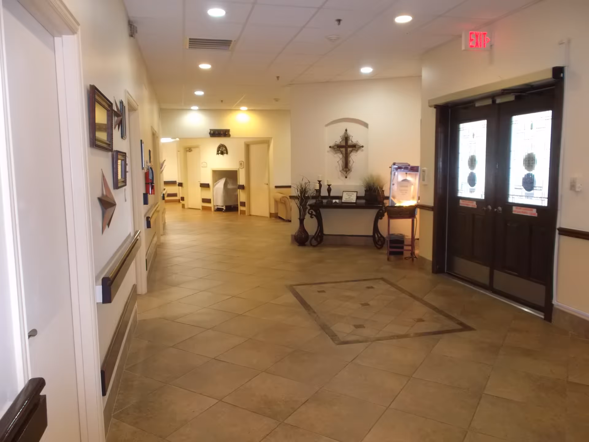 A well-lit hallway in a nursing and rehabilitation facility with beige tiled floors and cream-colored walls. The hallway features decorative wall art including framed pictures and a large cross above a console table with vases and candles. Double doors with stained glass panels and an exit sign are visible on the right side. The hallway has handrails along the walls and recessed ceiling lights.