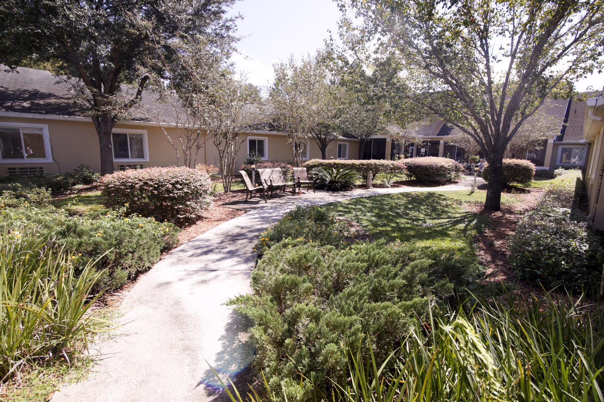 A sunny outdoor courtyard at Brookdale Avondale featuring a winding concrete pathway surrounded by green shrubs, bushes, and trees. Several benches are placed along the path for seating, and the building with beige walls and white-framed windows encloses the garden area.