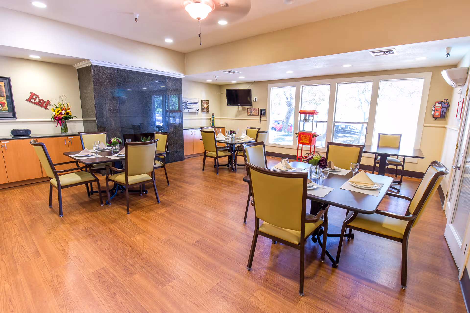 A bright dining room with wooden floors and several tables set with plates, glasses, and napkins. The room has large windows letting in natural light, yellow cushioned chairs, a popcorn machine near the windows, a wall-mounted TV, and a cabinet with a flower arrangement and a decorative 'Eat' sign on the wall.