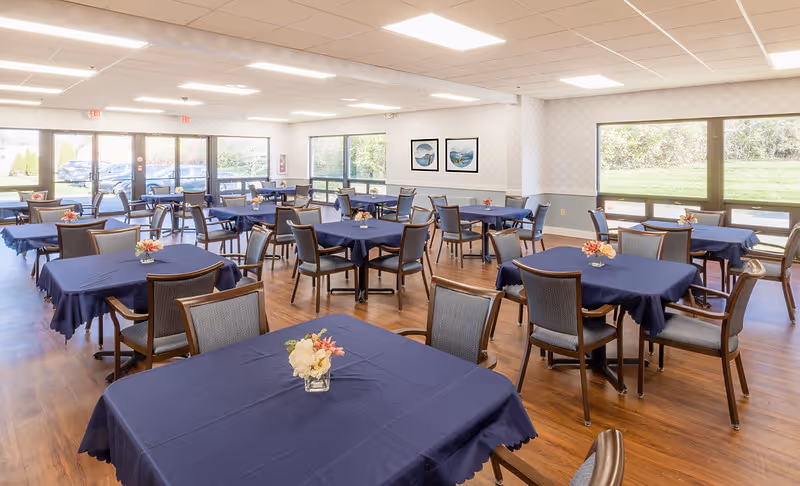 Spacious dining room with multiple tables draped in navy tablecloths, chairs, and small floral centerpieces beneath bright ceiling lights and large windows.