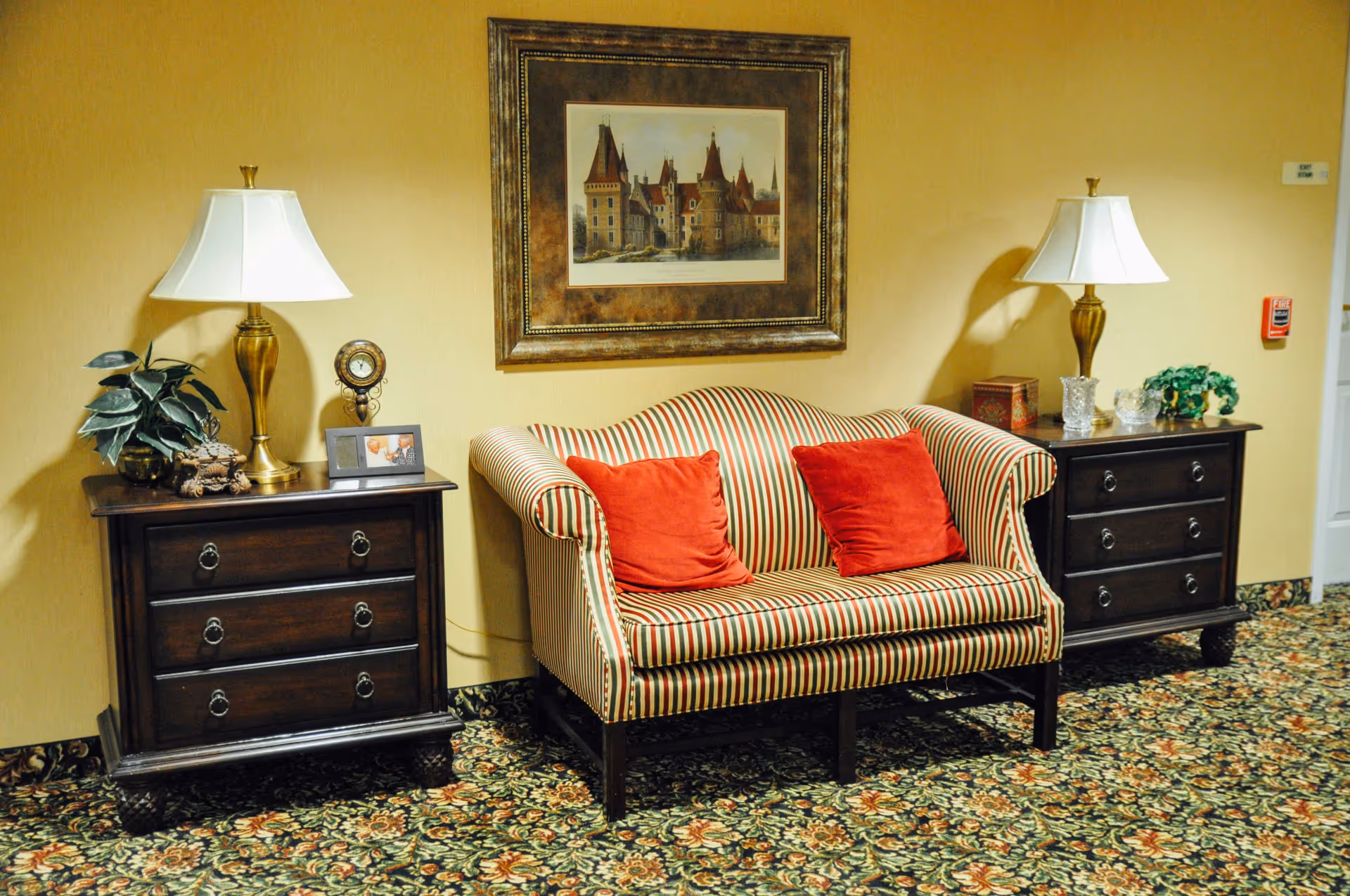 A cozy sitting area with a striped loveseat featuring red cushions, flanked by two dark wooden side tables each holding a brass lamp with a white shade. One side table has a small plant, a clock, and a framed photo, while the other has decorative items and a small green plant. Above the loveseat is a framed picture of a castle. The floor is covered with a floral patterned carpet and the walls are painted a warm beige color.