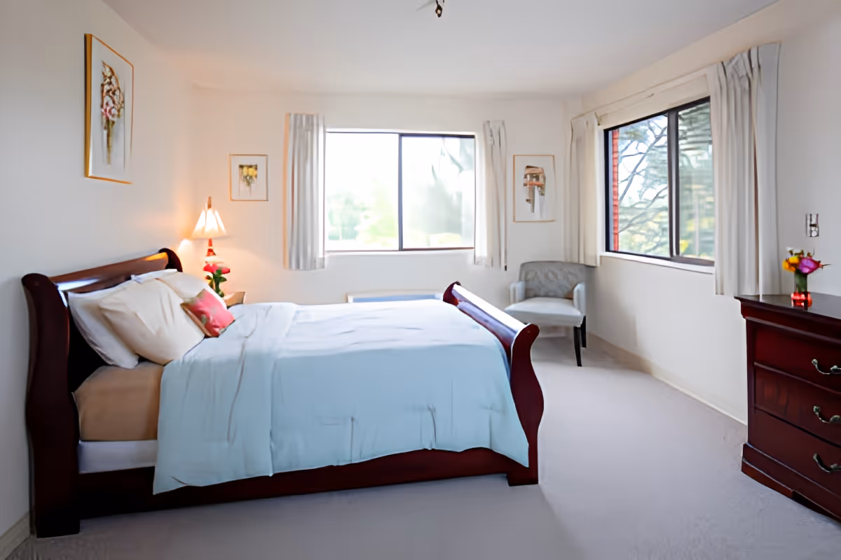 Bright, tidy bedroom with a wooden sleigh bed, two windows, a dresser and an upholstered chair.