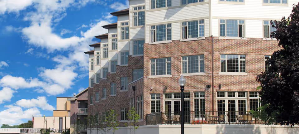Exterior view of a multi-story senior living facility building with a brick facade and numerous windows under a partly cloudy blue sky. There is a patio area with tables and chairs visible on the ground level, along with some trees and a streetlamp in front.
