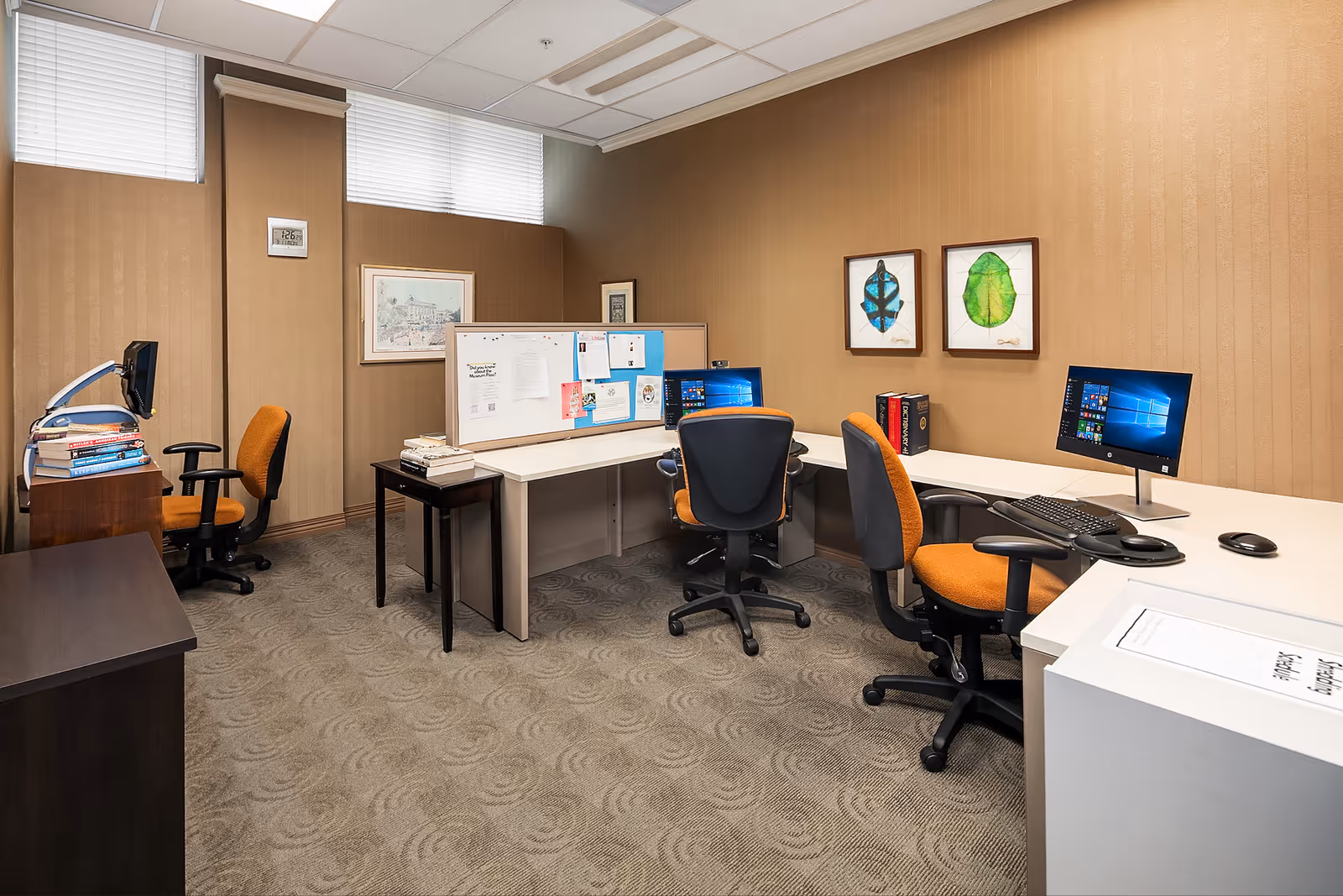 An office space with multiple desks and orange office chairs. There are computer monitors on the desks, a bulletin board with papers pinned on it, framed artwork on the walls, and a stack of books on one desk. The room has beige walls and carpeted flooring with a patterned design.