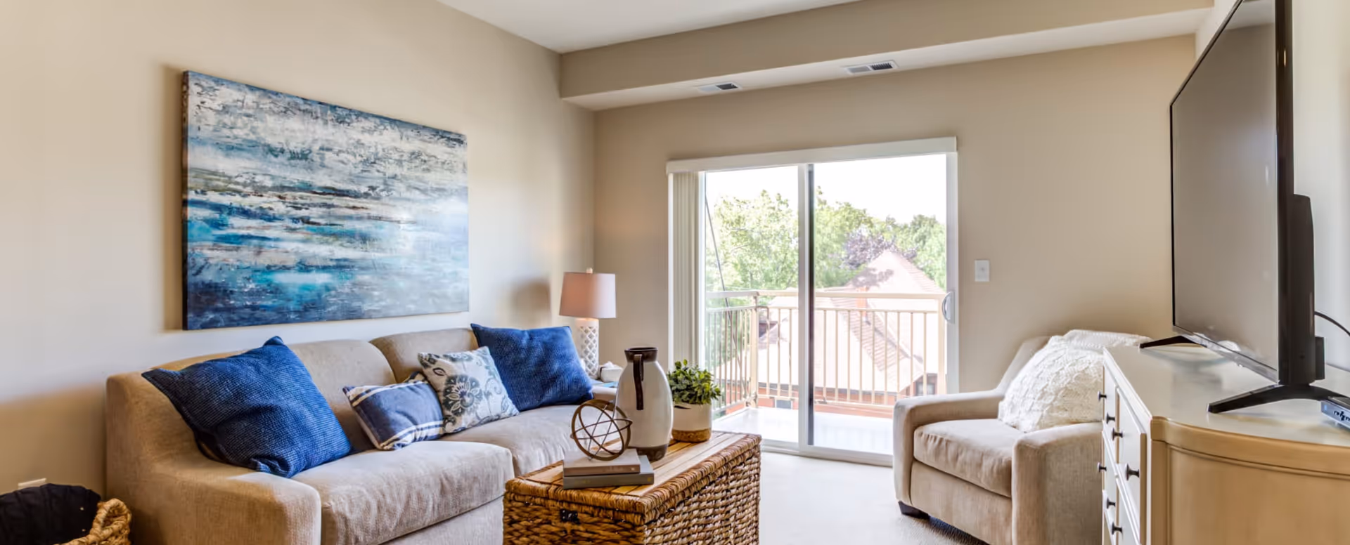 Bright living room with a beige sofa and armchair, blue accent pillows, wicker coffee table, wall art, and sliding glass doors opening to a balcony.