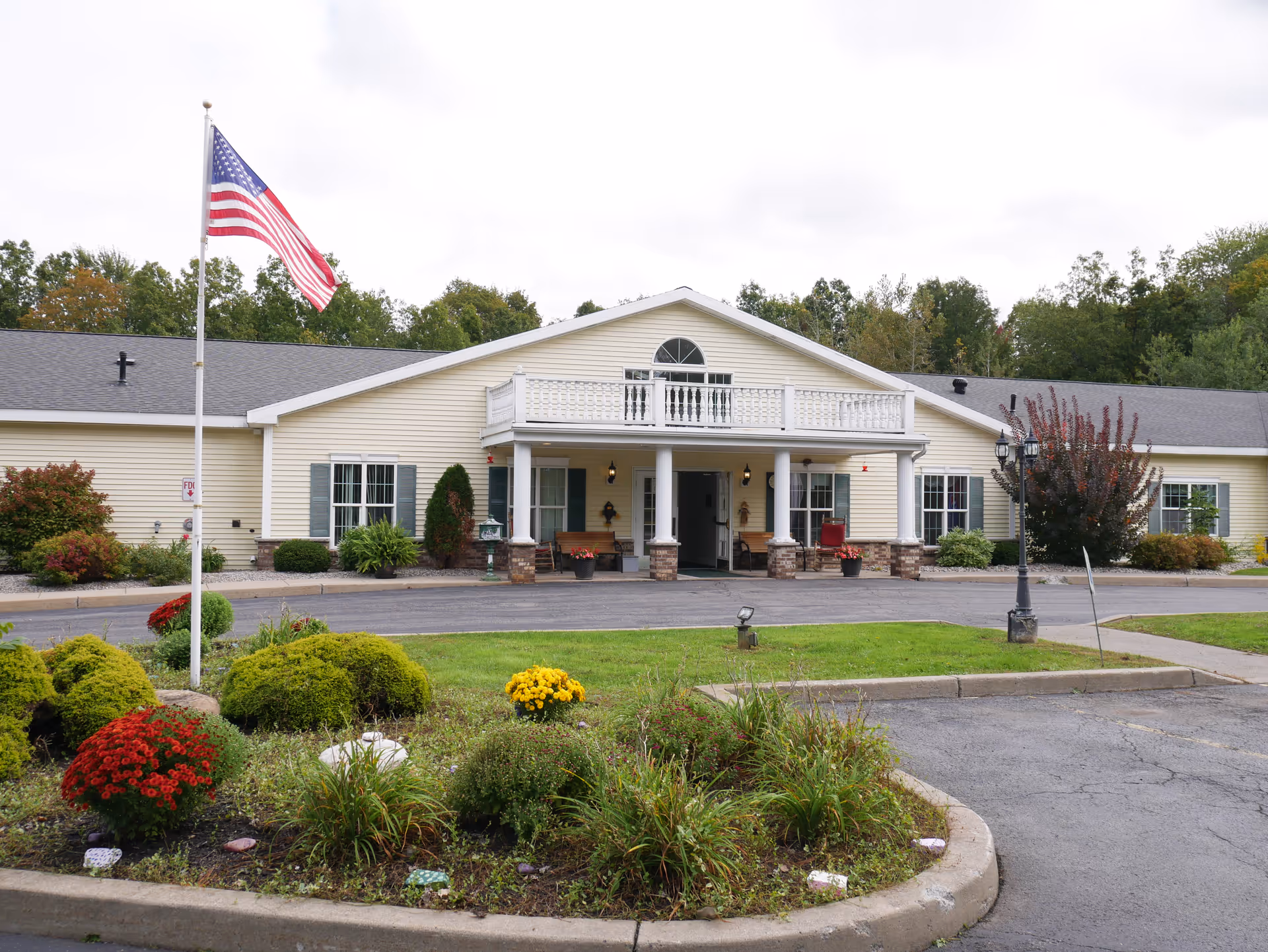Front exterior view of a single-story senior living facility building with beige siding, a covered entrance supported by white columns, and a balcony above the entrance. There is an American flag on a flagpole in the landscaped front yard with bushes and flowers. Trees are visible in the background under a cloudy sky.