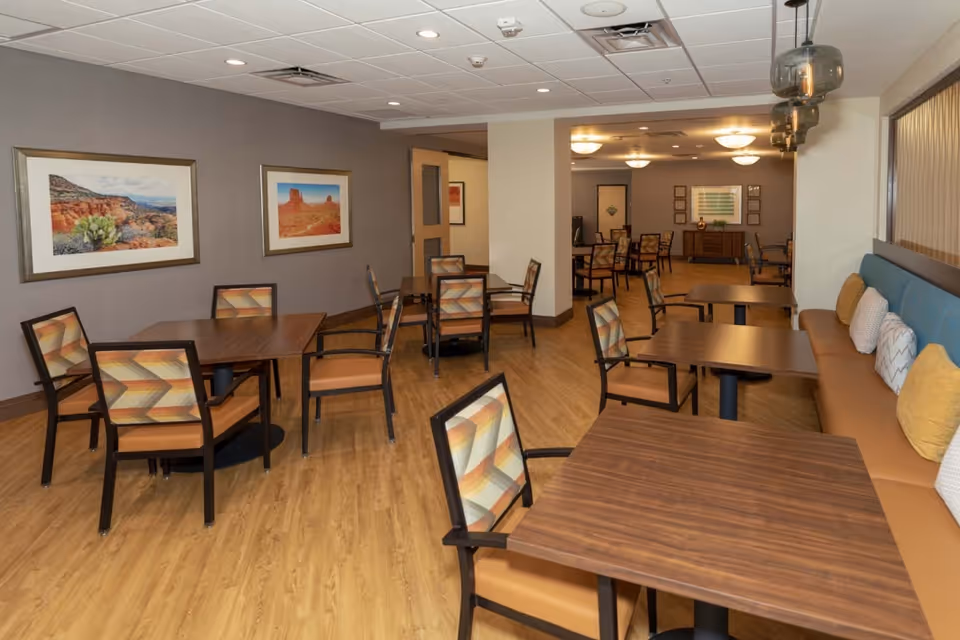 Interior view of a dining room in a senior living facility with wooden tables and chairs featuring patterned cushions. The room has wood flooring, framed landscape pictures on the wall, and a long cushioned bench with decorative pillows along one side. Ceiling lights and hanging pendant lights illuminate the space.