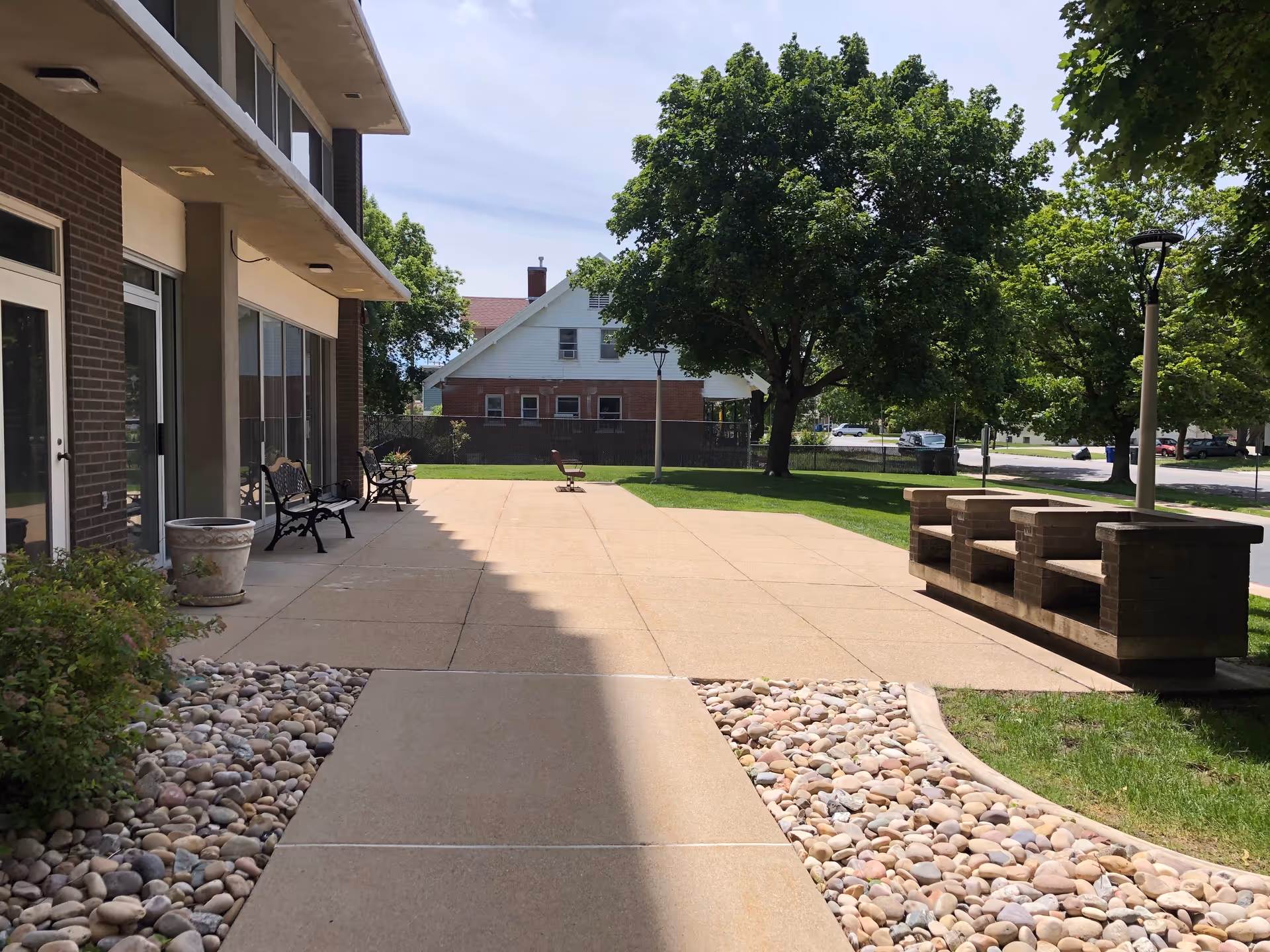 Outdoor patio area at Fellowship Manor with benches, a chair, a paved walkway, decorative rocks, green grass, large trees, and a residential house in the background under a clear sky.