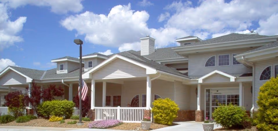 Front exterior of an assisted living facility with a covered entrance, columns, American flag, and landscaped beds under a blue sky.