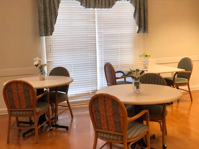 Small dining area with round tables and cushioned chairs arranged near a large window with blinds and vases of flowers on the tables.
