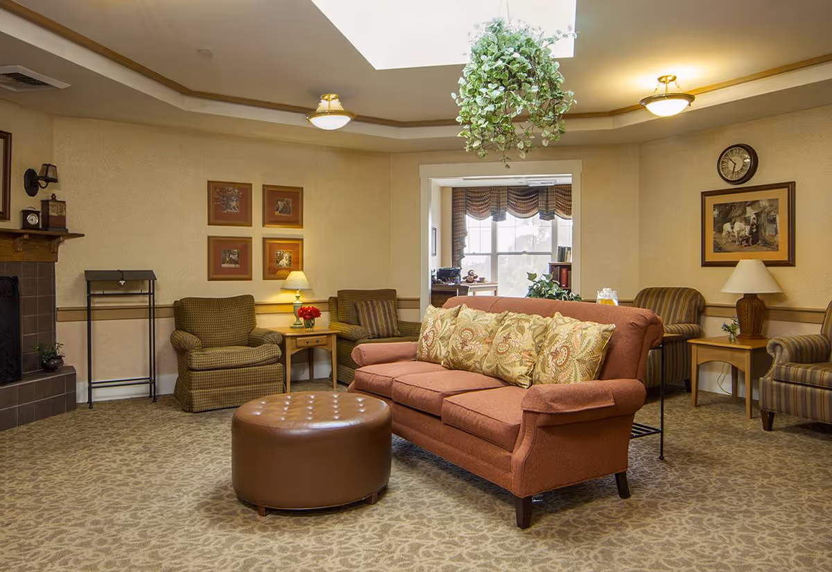 A cozy living room area with a brown sofa adorned with patterned cushions, a round brown ottoman, and several armchairs. The room features beige walls with framed artwork, a fireplace on the left, a hanging plant from the ceiling, and a window with valances letting in natural light. There are side tables with lamps and decorative items, and a clock on the wall.