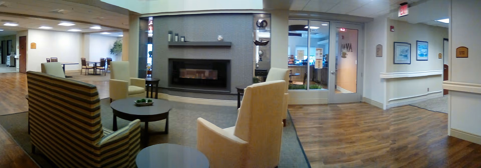Interior view of a senior living facility common area with comfortable seating including armchairs and a striped sofa around small round tables. A modern fireplace is built into a gray tiled wall. In the background, there are more tables and chairs, a hallway, and glass doors leading to another room. The floor is wood with some carpeted areas, and the walls are light-colored with handrails and framed pictures.