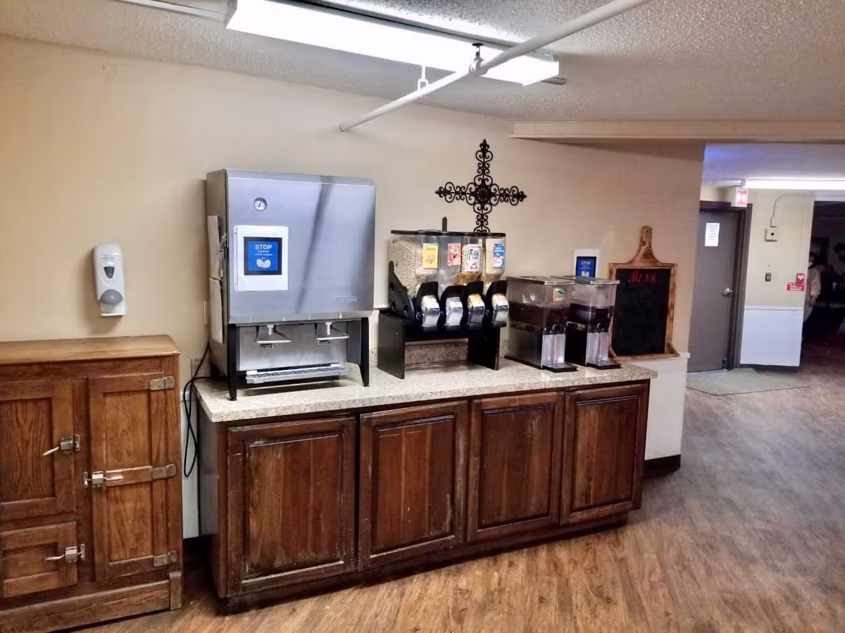 A beverage station in a hallway with a water dispenser, cereal dispensers, and two juice containers on a wooden cabinet. There is a wall-mounted hand sanitizer dispenser on the left and a decorative cross on the wall above the cereal dispensers. A small chalkboard menu is on the right side of the cabinet, and a hallway with doors is visible in the background.