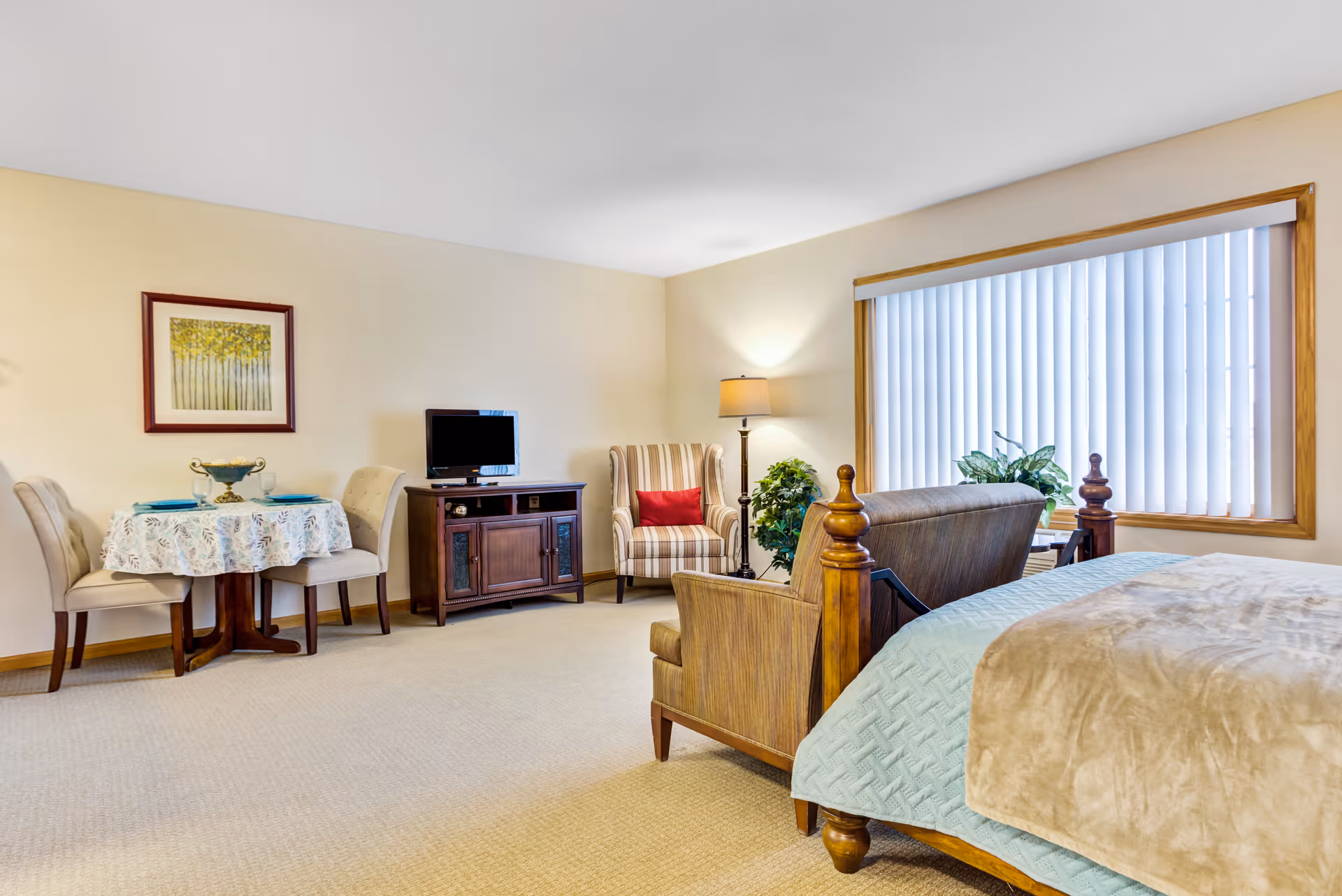 A cozy senior living room and bedroom area featuring a wooden bed with a light blue quilt and beige blanket, a striped armchair with a red pillow, a wooden TV stand with a small television, a round dining table covered with a floral tablecloth and set with two place settings, a floor lamp, and a large window with vertical blinds letting in natural light.