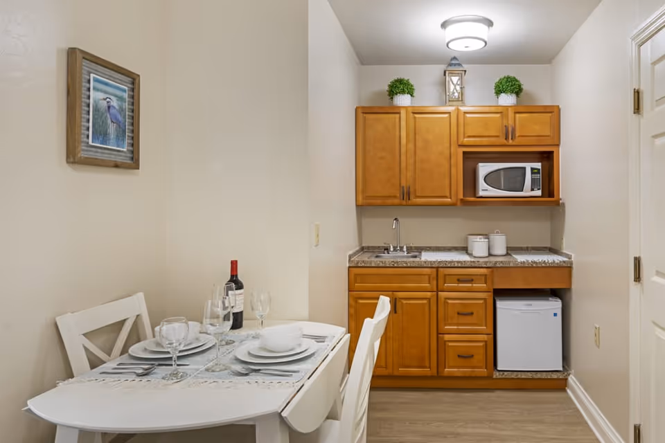 Small kitchenette area with wooden cabinets, a microwave, a mini refrigerator, a sink, and countertop with two canisters. Adjacent to the kitchenette is a small white dining table set for two with plates, bowls, wine glasses, and a bottle of wine. A framed picture of a bird hangs on the beige wall above the table.