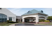 Front exterior view of La Bella of Edwardsville facility showing a covered entrance with a circular driveway, white walls, large windows, and a green metal roof under a partly cloudy sky.