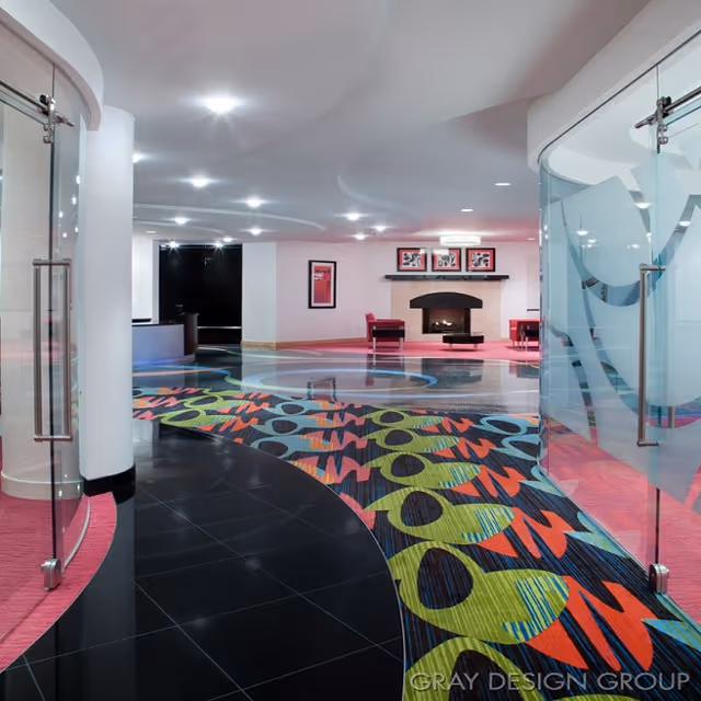 Modern brightly colored lobby with patterned carpet, glass doors, and a seating area with a fireplace.