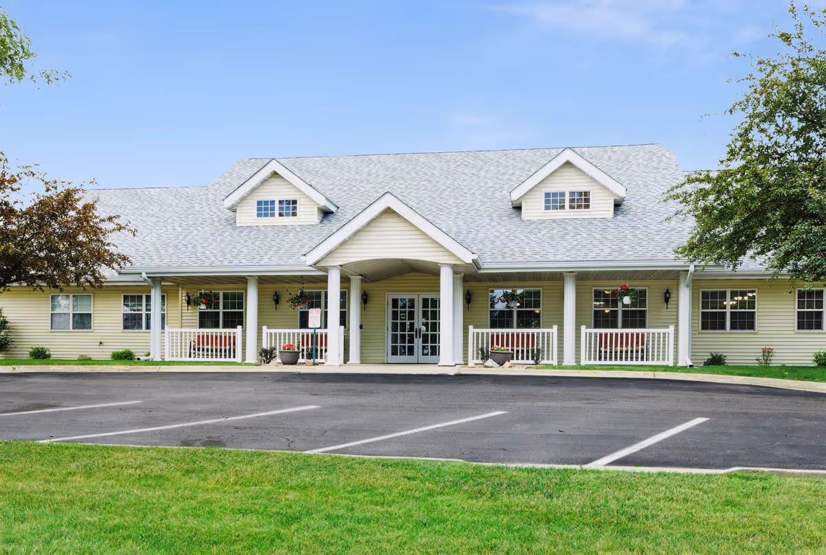 Front exterior view of a single-story senior living facility building with a light gray roof, beige siding, white columns, and a covered entrance. There are hanging flower baskets and potted plants near the entrance, with a paved parking area and green lawn in the foreground under a clear blue sky.