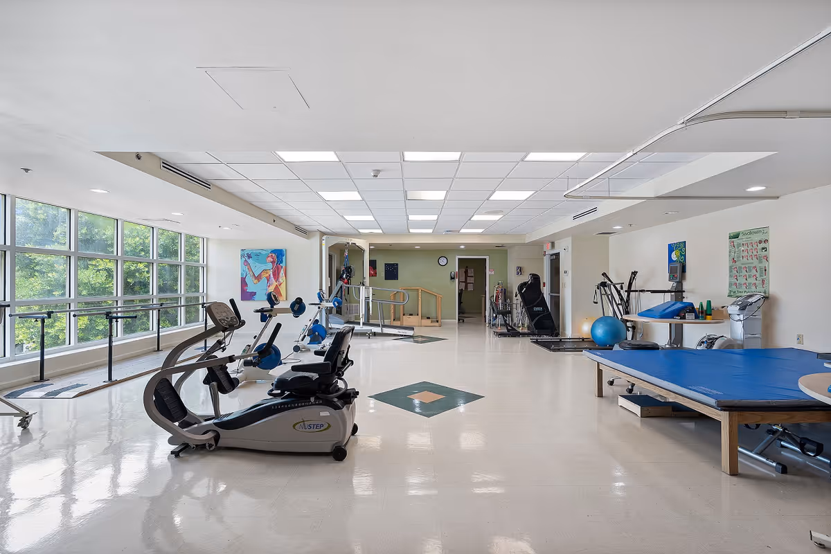 A spacious rehabilitation or physical therapy room with exercise equipment including stationary bikes, parallel bars, therapy tables, and various exercise tools. Large windows on the left side allow natural light to fill the room, and colorful artwork is displayed on the walls.