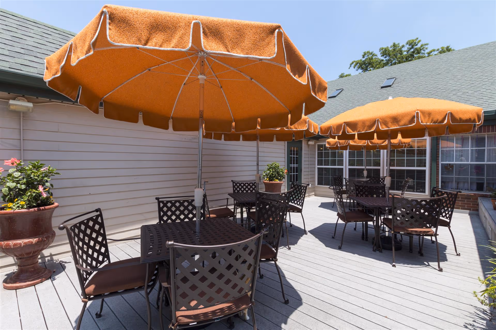 Outdoor patio area with multiple tables and chairs under large orange umbrellas. There are potted plants placed around the deck, and the building exterior with windows and a door is visible in the background under a clear blue sky.