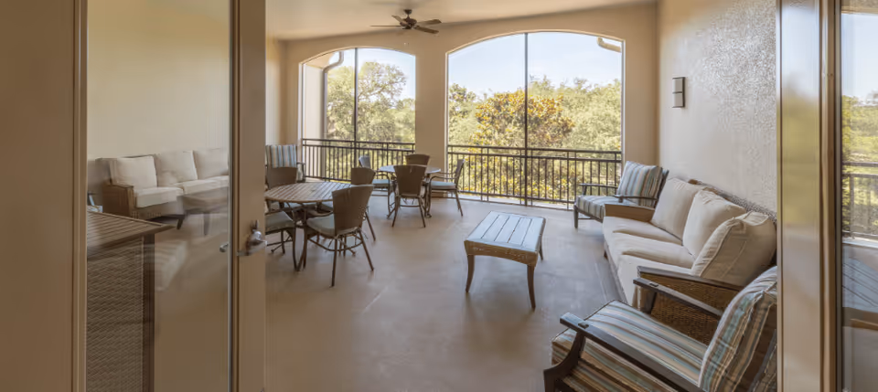 A spacious indoor lounge area with large windows overlooking trees. The room features cushioned wicker sofas, armchairs, a coffee table, and round tables with chairs, creating a comfortable sitting and socializing space.