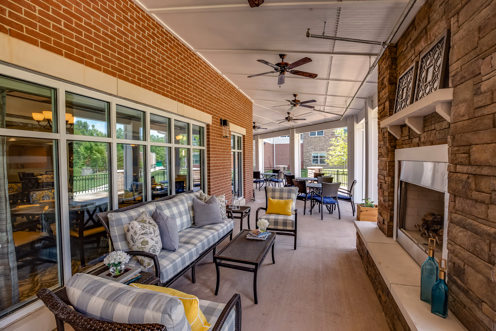 Covered outdoor patio with cushioned seating, tables, ceiling fans and a stone fireplace alongside large windows.