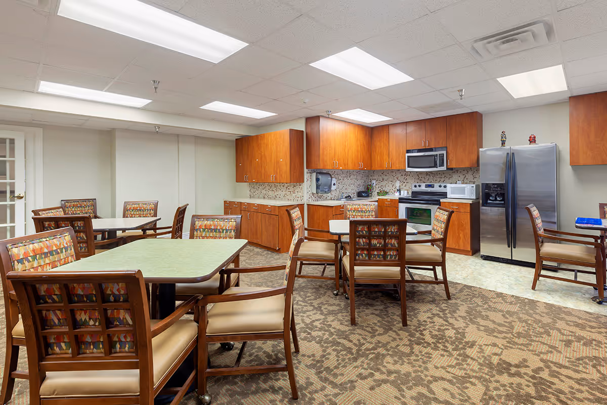 A communal dining and kitchen area in an assisted living facility featuring multiple tables with wooden chairs that have patterned upholstery. The kitchen area includes wooden cabinets, a stainless steel refrigerator, a microwave, an oven, and a countertop with a mosaic tile backsplash. The room is well-lit with ceiling lights and has a carpeted floor with a patterned design.